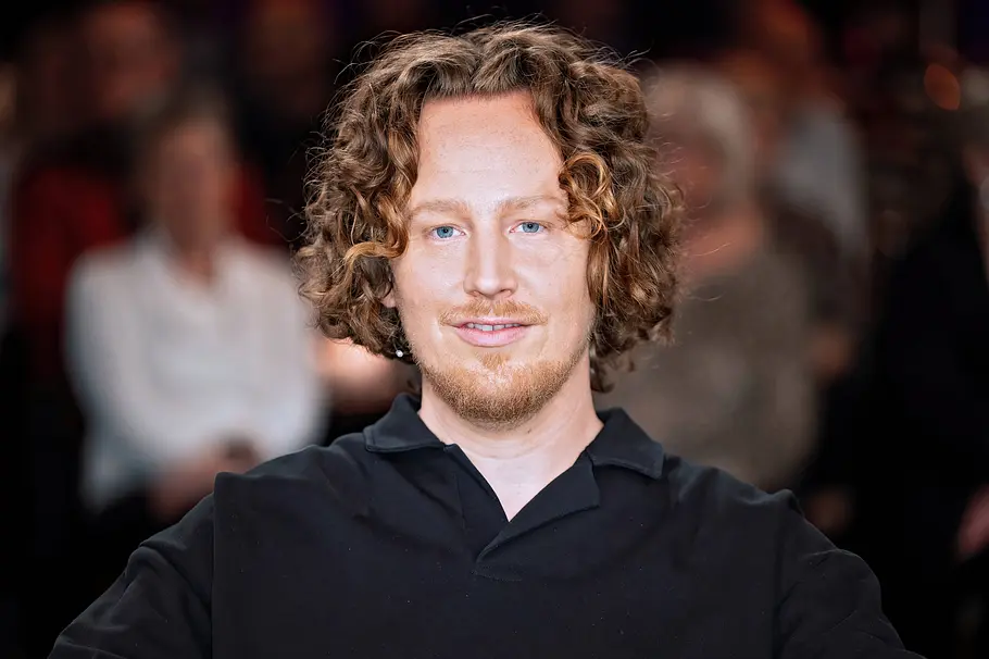 „The Voice“ Gast-Coach Michael Schulte. Michael Schulte mit lockigem Haar und Bart trägt ein schwarzes Poloshirt. Hinter ihm sitzen unscharf erkennbare Personen.
