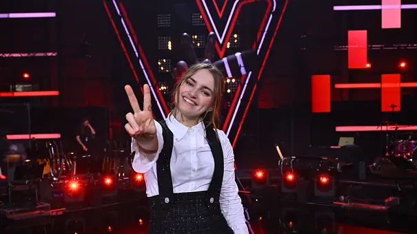Kandidatin Anne Mosters zeigt Peace in die Kamera. Sie trägt eine weiße Bluse, darüber einen schwarzen Overall. - Foto: Seven.One Entertainment Group GmbH Kandidatin Anne Mosters zeigt Peace in die Kamera. Sie trägt eine weiße Bluse, darüber einen schwarzen Overall. - Foto: Seven.One Entertainment Group GmbH