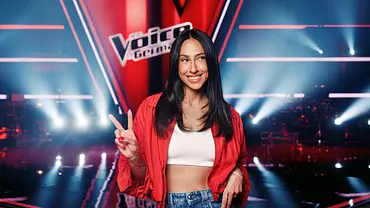 Melissa Khalaj lächelt in roter Jacke, weißem Top und Jeans vor der „The Voice“-Kulisse und zeigt das Peace-Zeichen. - Foto: Joyn