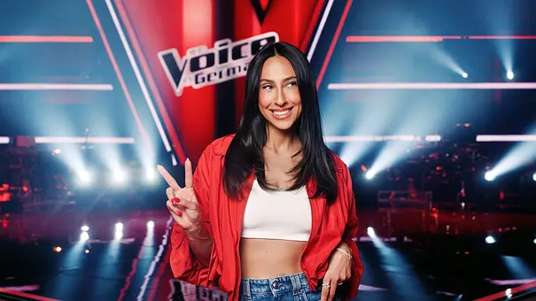 Melissa Khalaj lächelt in roter Jacke, weißem Top und Jeans vor der „The Voice“-Kulisse und zeigt das Peace-Zeichen. - Foto: Joyn Melissa Khalaj lächelt in roter Jacke, weißem Top und Jeans vor der „The Voice“-Kulisse und zeigt das Peace-Zeichen. - Foto: Joyn