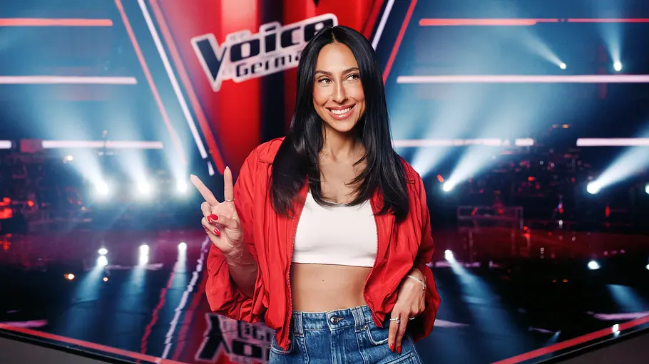 The Voice: Moderatorin Melissa Khalaj Melissa Khalaj lächelt in roter Jacke, weißem Top und Jeans vor der „The Voice“-Kulisse und zeigt das Peace-Zeichen.