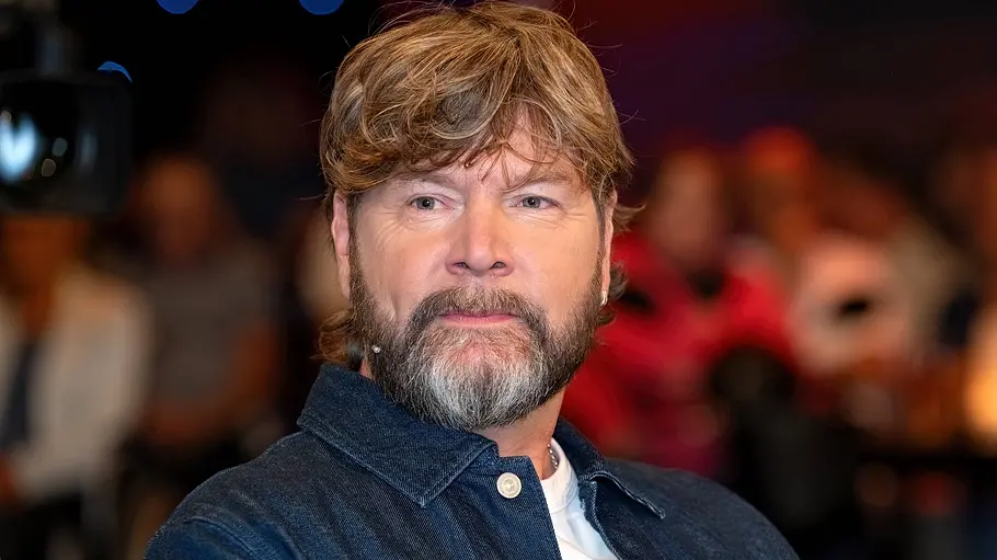 „The Voice of Germany“: Rea Garvey will beide Talente mitnehmen – doch eine steigt freiwillig aus! Rea Garvey trägt eine dunkle Jeansjacke über einem weißen Shirt. Im Hintergrund sind unscharf mehrere Personen zu sehen.