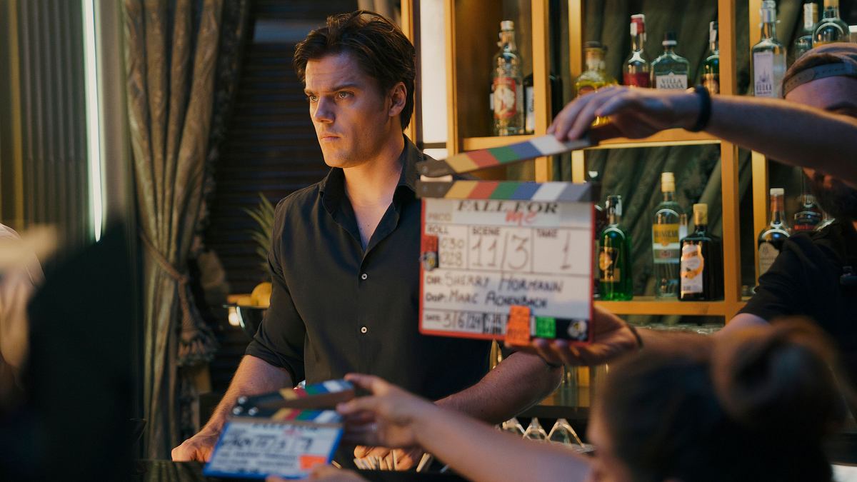 Theo Trebs steht in seiner Rolle als Tom an einer Bar. Die Filmcrew hält die Klappe in die Kamera, während hinter ihm eine Regalwand voller Flaschen zu sehen ist.