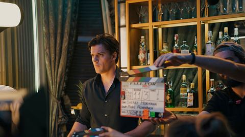 Theo Trebs steht in seiner Rolle als Tom an einer Bar. Die Filmcrew hält die Klappe in die Kamera, während hinter ihm eine Regalwand voller Flaschen zu sehen ist. - Foto: Julia Terjung/Netflix