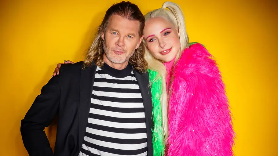 Theresia Fischer und Stefan Kleiser Theresia Fischer im pink-grünen Zottelmantel und Stefan Kleiser im schwarzen Sakko stehen eng zusammen vor einer gelben Wand.