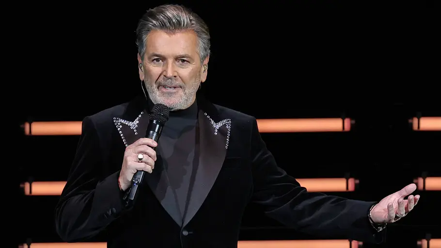 Thomas Anders ZDF Weihnachtsshow Thomas Anders hält ein Mikrofon und streckt einen Arm aus. Er trägt ein schwarzes Sakko vor orangefarbenen Bühnenlichtern.