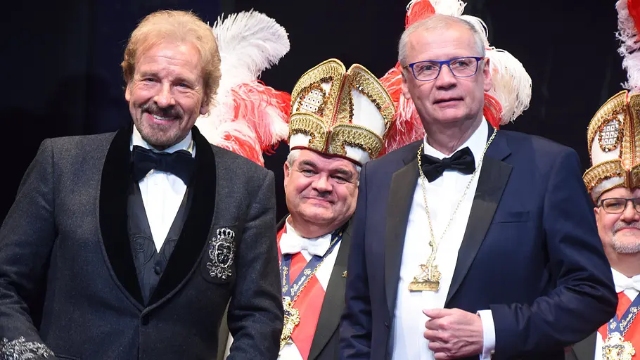 Man sieht Gottschalk und Jauch in zwei Anzügen auf einer Art Karnevalsveranstaltung. Hinter ihnen stehen Personen in entsprechenden Aufzügen.