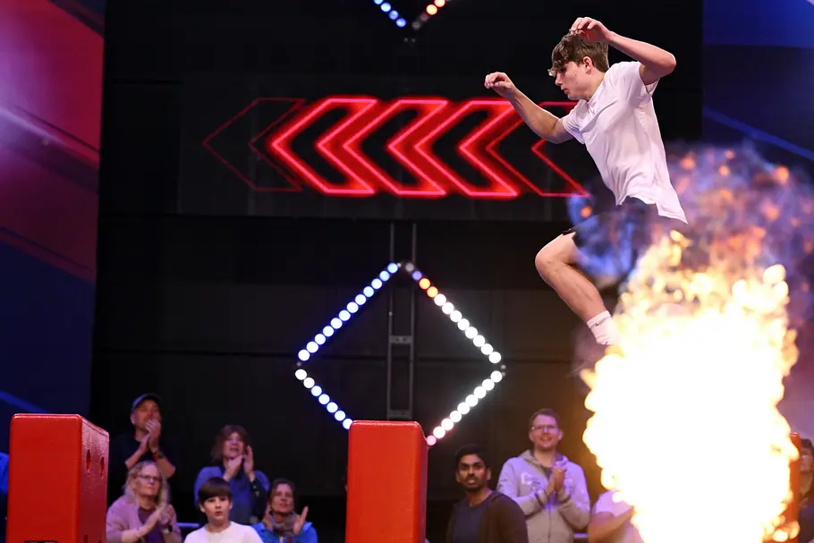 Thomas Grandl ist Teilnehmer bei Ninja Warrior Germany 2026 Thomas Grandl springt mit erhobenen Armen über rote Hindernisse. Im Vordergrund lodert eine Flamme, dahinter sitzt Publikum.