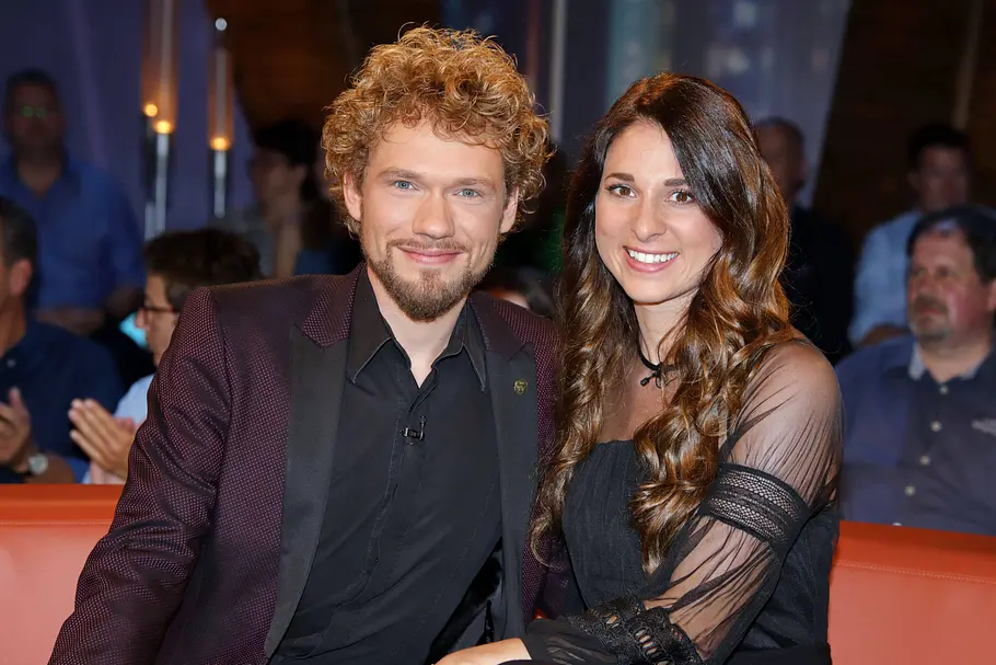 Thommy Ten und Amélie van Tass alias The Clairvoyants stzen auf einer orangenen Couch und lächeln gemeinsam in die Kamera
