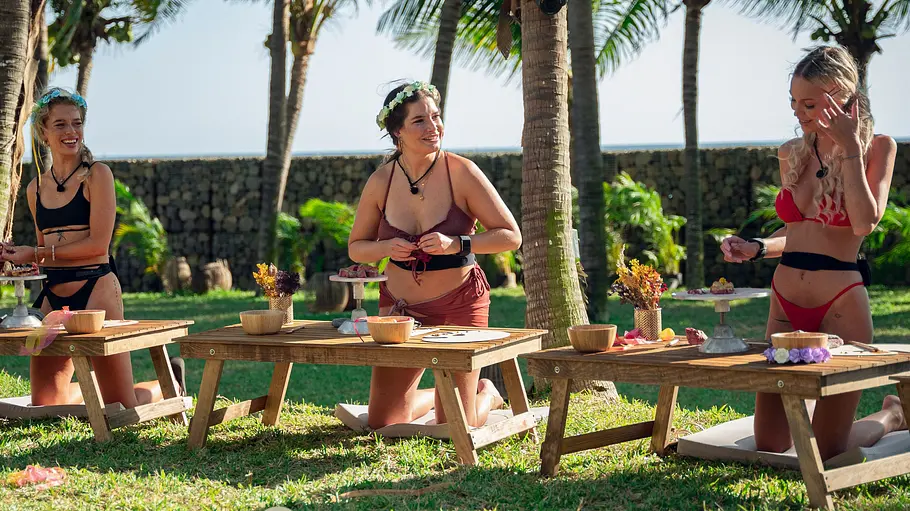 Laura Sophie Gehrke in „Too Hot To Handle: Germany“ Emely Hüffer, Laura und Laura Sophie Gehrke knien in Staffel 1 von Too Hot to Handle: Germany in Bikinis auf einer Wiese unter Palmen.