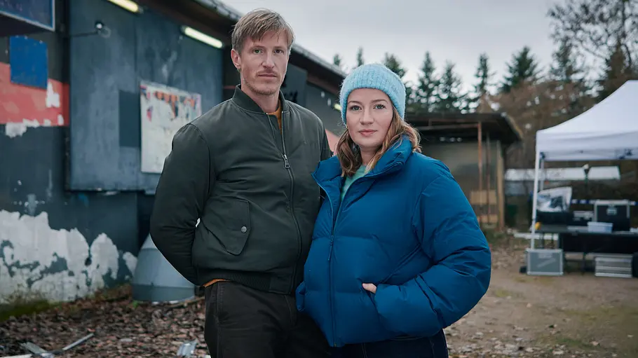 Thüringenkrimi – CASH (AT): Moritz Herbst (Golo Euler) und Theresa Wolff (Nina Gummich) stehen mit den Händen in den Taschen vor einer Holzhütte und blicken in die Kamera.