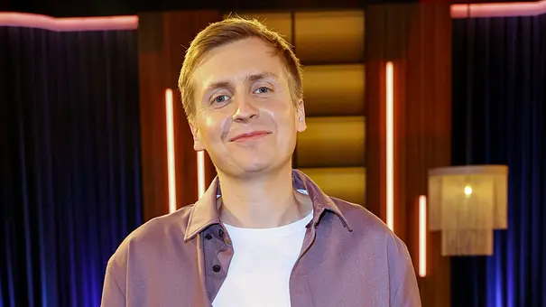 Till Reiners lächelnd in einem TV-Studio mit warm beleuchtetem Hintergrund und vertikalen Lichtstreifen - Foto: IMAGO / BREUEL-BILD