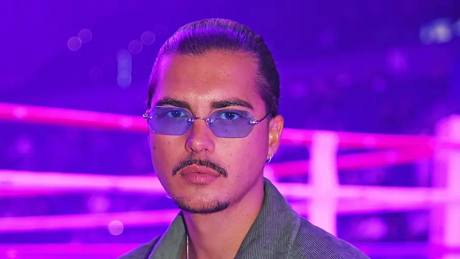 Tim Kühnel Tim Kühnel steht vor einem Boxring, der in lila Licht getaucht ist. Er trägt eine durchsichtige Sonnenbrille und eine grüne Cordjacke und schaut ernst in die Kamera.