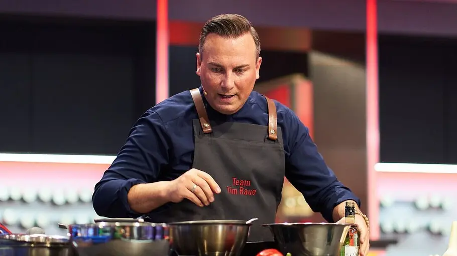 Tim Raue The Taste Tim Raue steht in dunkler Kochjacke und Schürze hinter einer Kochtheke mit Metallschüsseln, Paprika, Zucchini und Knoblauch.