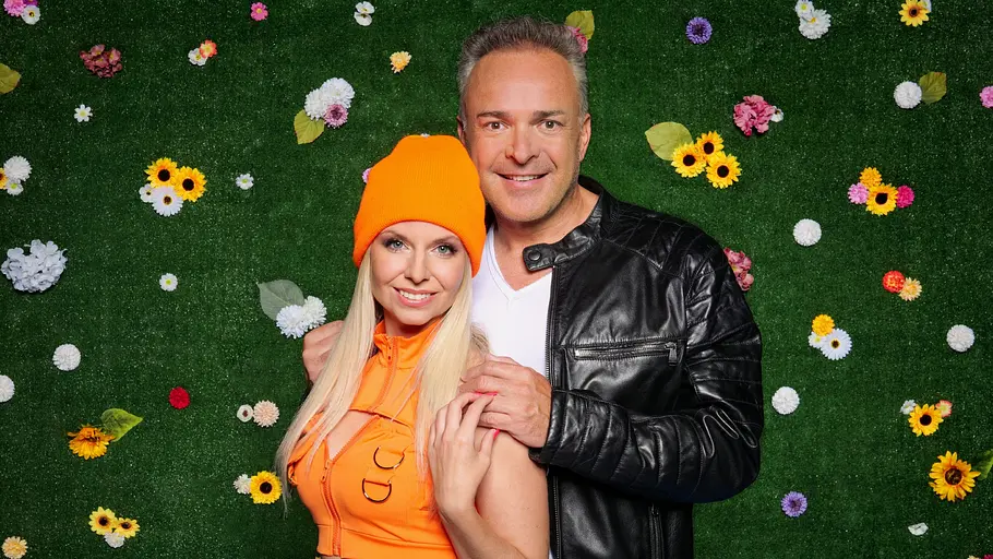 Tim Toupet und Carina Crone Tim Toupet in schwarzer Lederjacke und Carina Crone mit orangefarbener Mütze und Top posieren vor einer grünen Blumenwand.