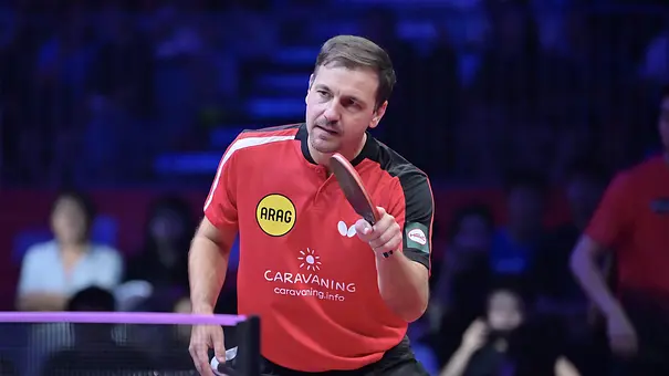Timo Boll im roten Trikot hält seinen Schläger über dem Tisch. Im Hintergrund sitzen unscharf erkennbare Zuschauer. - Foto: Joyn/Douyin Timo Boll im roten Trikot hält seinen Schläger über dem Tisch. Im Hintergrund sitzen unscharf erkennbare Zuschauer. - Foto: Joyn/Douyin