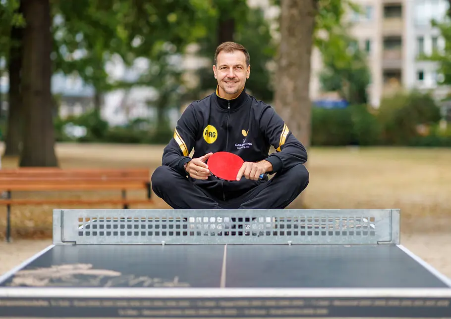 Timo Boll im Trainingsanzug sitzt im Schneidersitz auf einer Tischtennisplatte im Park und hält einen roten Schläger.