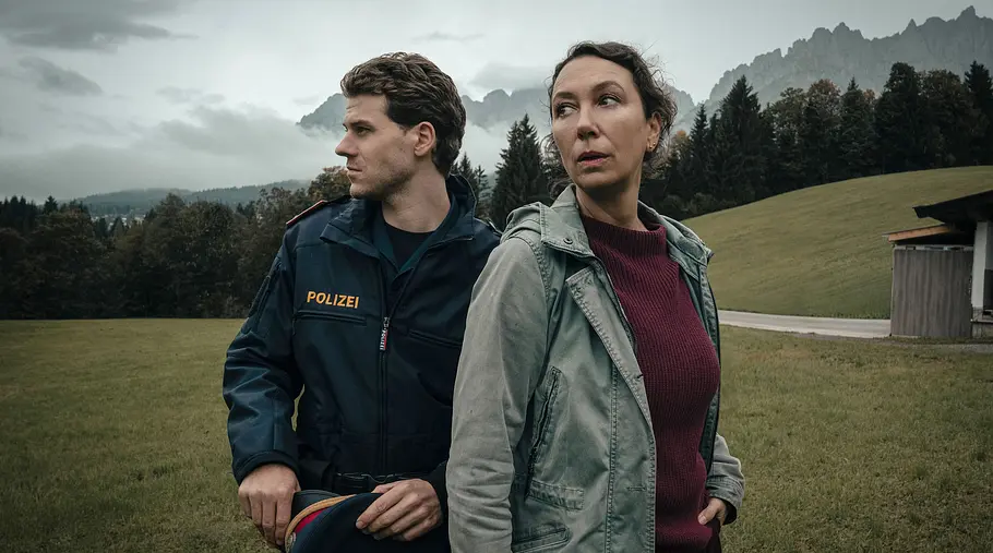 Tiroler Blut: Der erste Schuss: Chefinspektorin Fanny Gruber (Ursula Strauss) und Sohn Tobi (Noah L. Perktold) in Polizeiuniform stehen auf einer Wiese vor bewaldeten Bergen unter grauem Himmel.