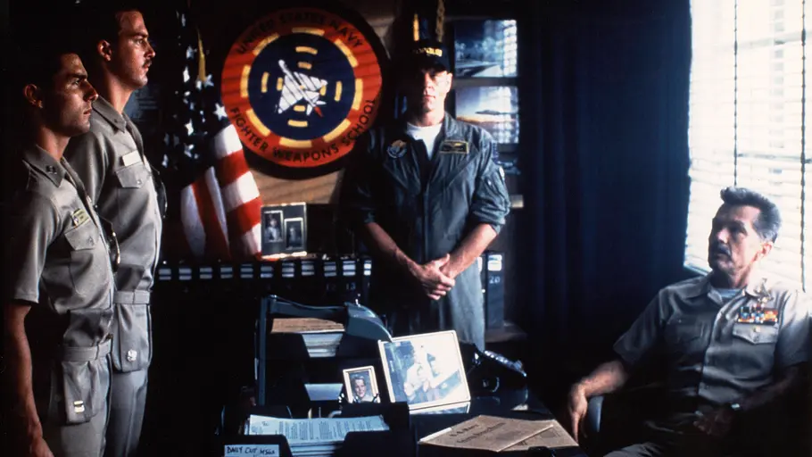 Tom Cruise und Tom Skerritt in „Top Gun“. Drei Männer in Uniform stehen vor einem sitzenden Offizier am Schreibtisch. Dahinter hängen eine US-Flagge und ein Navy-Abzeichen.