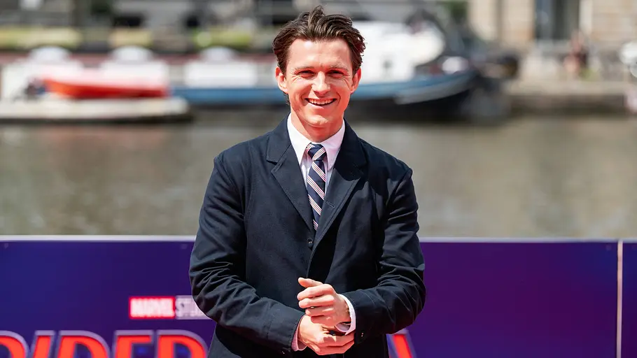 Tom Holland Ein Mann in dunklem Anzug, weißem Hemd und gestreifter Krawatte steht lächelnd auf einem roten Teppich vor einer Werbewand zum Film „Spider-Man“. Im Hintergrund sind ein Gewässer, Boote und Gebäude zu sehen. Die Aufnahme entstand bei Tageslicht und zeigt den Schauspieler bei einem öffentlichen Filmpremieren- oder Presseevent.