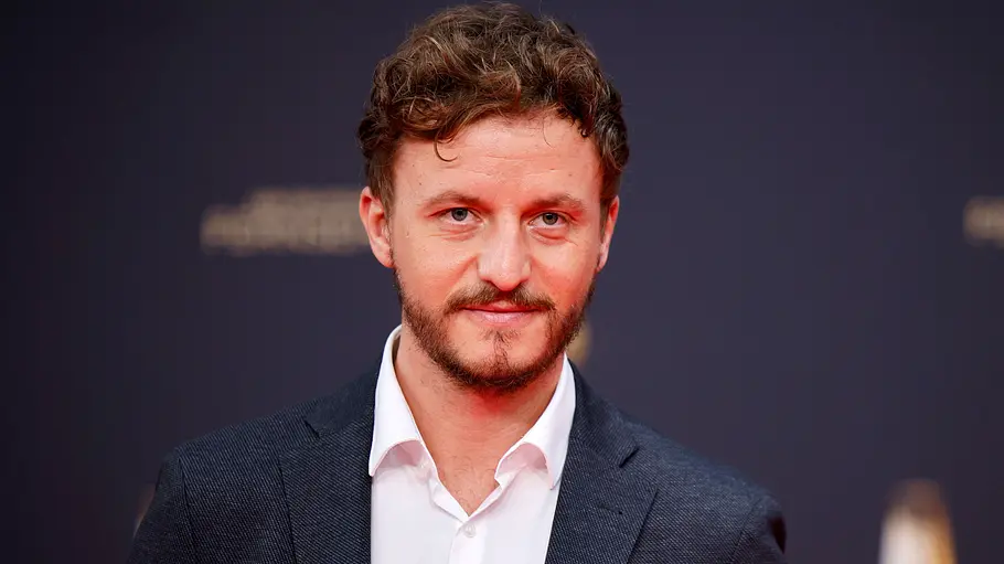 Tommi Schmitt trägt ein dunkles Sakko mit weißem Hemd und Einstecktuch und blickt vor dunklem Hintergrund in die Kamera.