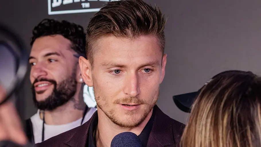 Tommy Pedroni Tommy Pedroni steht im dunklen Sakko vor einem Mikrofon. Eine Frau und eine Kamera sind im Vordergrund, ein Mann hinter ihm.