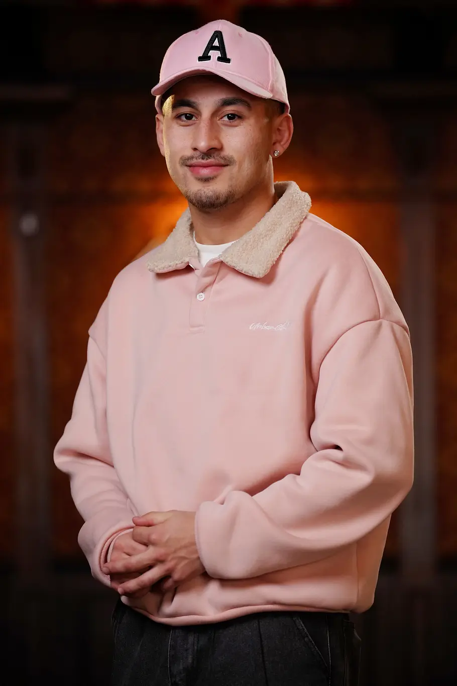 Abii Faizan Abii mit rosafarbener Cap und passendem Sweatshirt steht mit verschränkten Händen vor warm beleuchtetem Hintergrund.