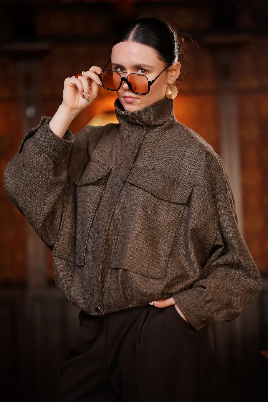 Constance Dizendorf Constanze mit zurückgebundenen Haaren trägt eine braune Jacke und hebt eine orange getönte Sonnenbrille leicht an.