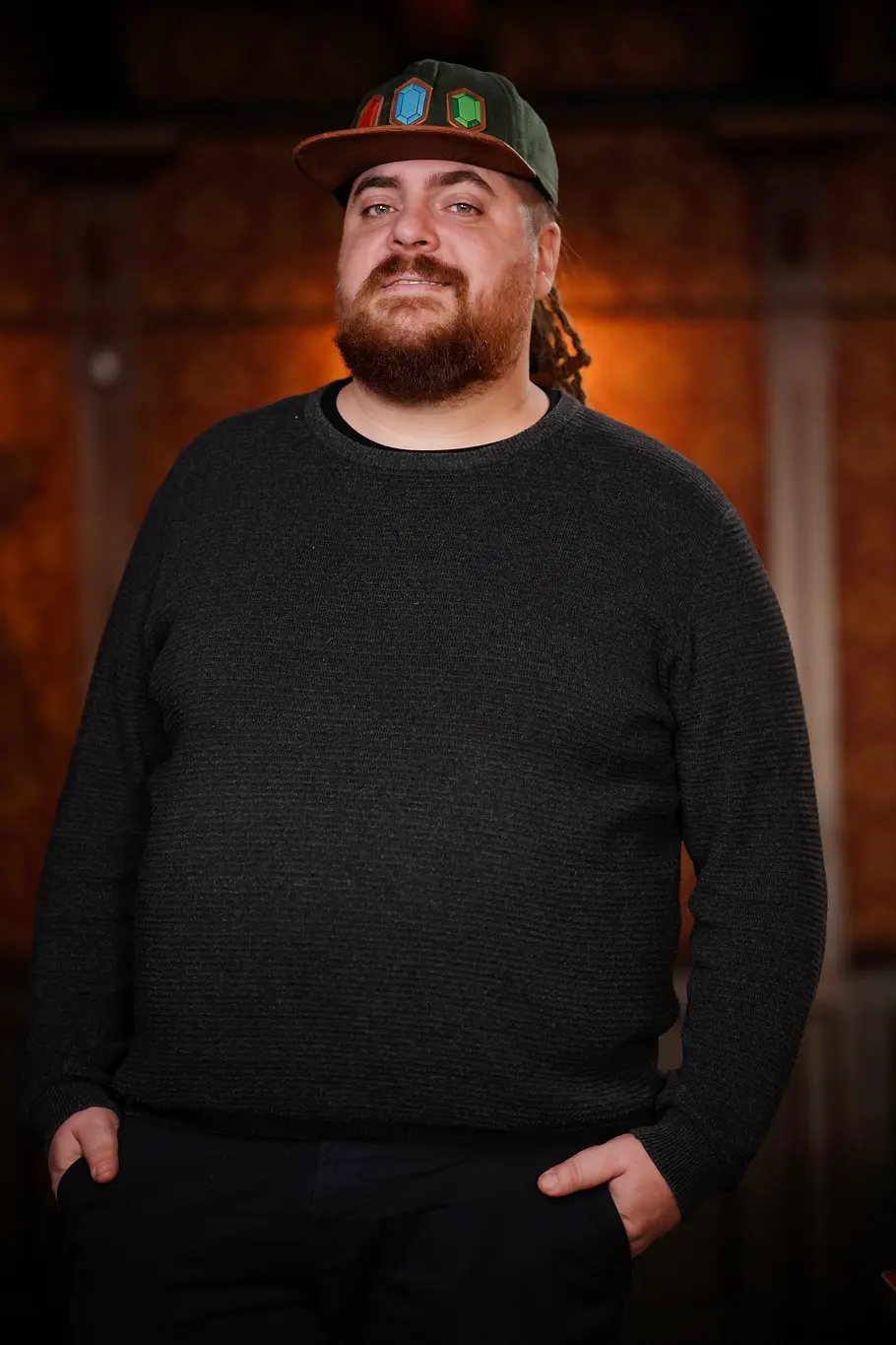 Lex Spielhaupter Lex mit Bart und dunklem Pullover trägt eine schwarze Cap mit farbigen Symbolen und steht vor warm beleuchtetem Hintergrund.