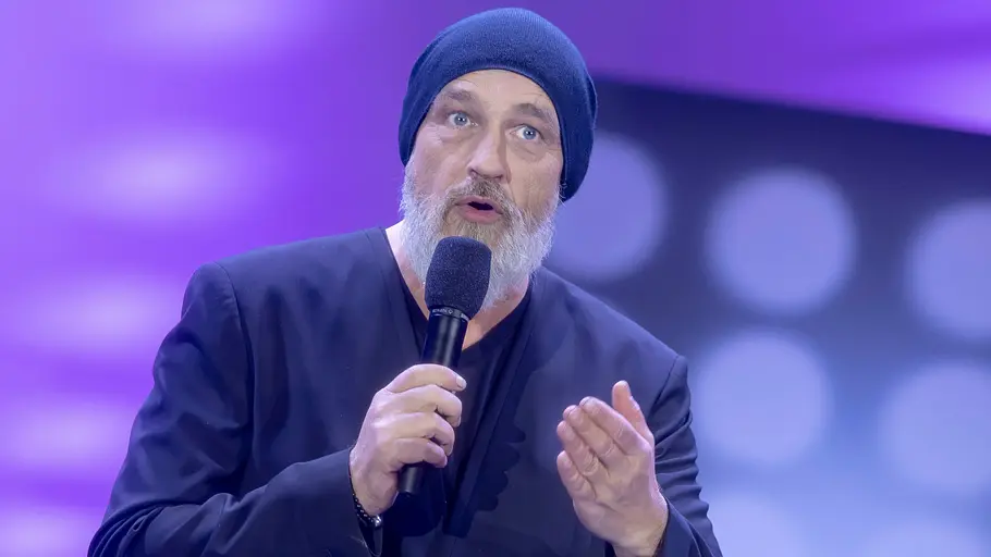 Torsten Sträter Torsten Sträter mit grauem Bart, dunklem Sakko und Mütze spricht mit einem Mikrofon auf einer Bühne mit lila Hintergrund.