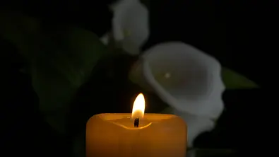 Eine Kerze leuchtet, im Hintergrund eine weiße Blume. - Foto: IMAGO / Zoonar