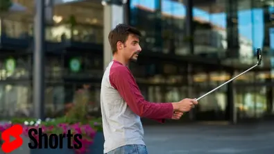 Mann mit Handy und Selfiestick vor Gebäude. YouTube Shorts Logo unten links. - Foto: TV Movie / Canva / YouTube Shorts Mann mit Handy und Selfiestick vor Gebäude. YouTube Shorts Logo unten links. - Foto: TV Movie / Canva / YouTube Shorts