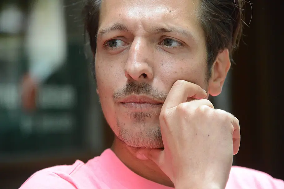 Thilo Mischke trägt ein rosa Shirt, stützt sein Kinn auf eine Hand und blickt nach links. Sein Gesicht ist in Nahaufnahme zu sehen.