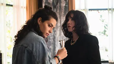 Alba hält einen Champagner in ihrer rechten Hand und schaut zur Seite. Beátrice steht ihr gegenüber und schaut mit erschrockener Miene nach Vorne.  - Foto: Marie Genin / Netflix
