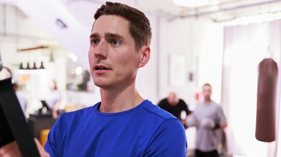 Ringo Ringo hält im blauen Sportshirt Trainingsgurte. Im hellen Fitnessraum stehen weitere Personen, rechts hängt ein Boxsack.