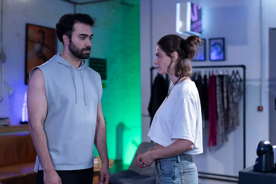 Unter Uns-Vorschau Freitag, 9.10.26, Folge 7974 Unter-Uns-Vorschau Freitag: Baris (Metehan Demiröz) im grauen, ärmellosen Hoodie und , Larissa (Ilka Mahrholz) im weißen T-Shirt stehen sich in einem grün und blau beleuchteten Raum gegenüber.