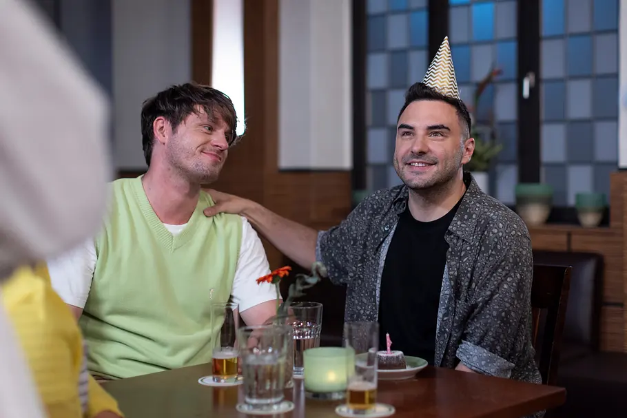Unter Uns-Vorschau heute, Montag, 5.10.26, Folge 7970 Unter Uns, Folge 7970: Joshua (Richard Lingscheidt, l.) sitzt lächelnd neben Easy (Lars Steinhöfel) am Tisch. Easy trägt einen Partyhut und legt Joshua eine Hand auf die Schulter.