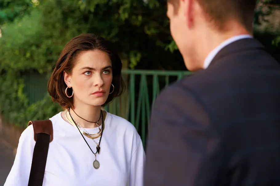 Unter Uns-Vorschau Mittwoch, 7.10.26, Folge 7972 Larissa (Ilka Mahrholz) schaut Ringo (Timothy Boldt) böse an. Sie trägt ein weißes Shirt und mehrere Ketten, er steht im dunklen Anzug mit dem Rücken zur Kamera.