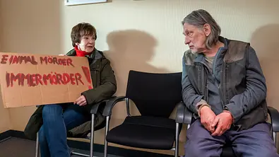 Katrin Sass hält ein Schild „Einmal Mörder, immer Mörder!“. Neben einem leeren Stuhl sitzt Wolfgang Michael mit Handschellen. - Foto: (S2+). NDR Presse und Information