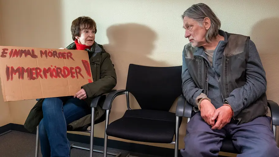 Usedom-Krimi: „Mörder unter uns“ Katrin Sass hält ein Schild „Einmal Mörder, immer Mörder!“. Neben einem leeren Stuhl sitzt Wolfgang Michael mit Handschellen.