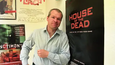 Uwe Boll posiert lächelnd neben einem Plakat seines Films „House of the Dead“. - Foto:  IMAGO / sepp spiegl