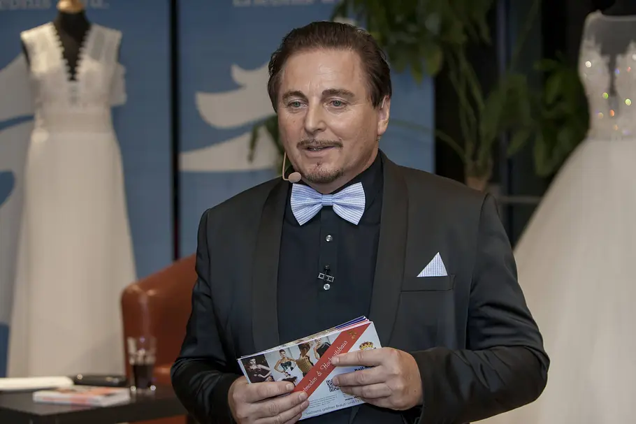 Uwe Herrmann von Tüll und Tränen Der Dresdner Modemacher Uwe Herrmann hält sein Buch in der Hand, im Hintergrund ein Tüll und Tränen-Brautkleid.