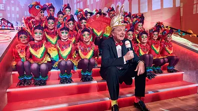 Moderator Uwe Koch präsentiert Westfalen haut auf die Pauke und sitzt inmitten der farbenfroh kostümierten Tänzerinnen und Tänzer im Congress Saal der Halle Münsterland. - Foto: © WDR 2026