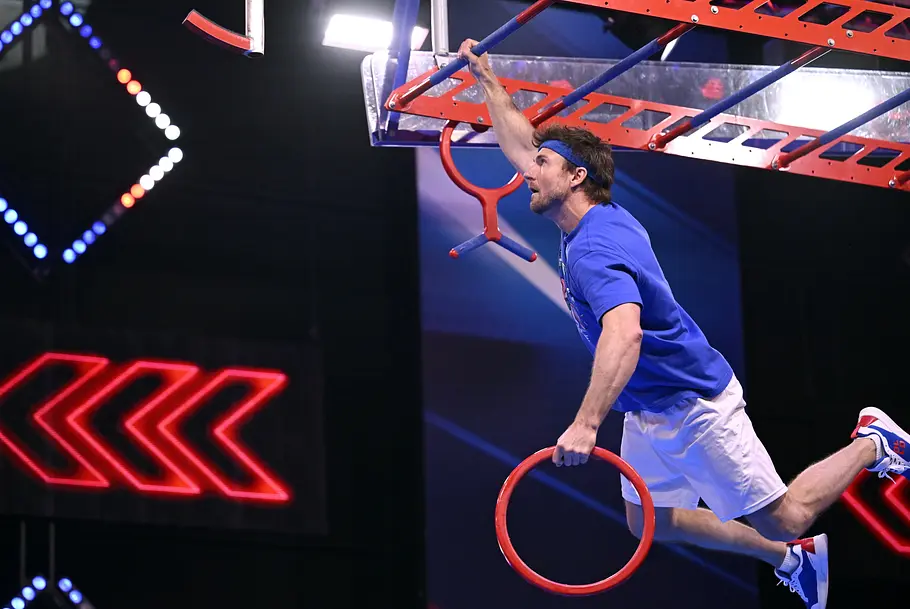 Uwe Weitzer ist Teilnehmer bei Ninja Warrior Germany 2026 Uwe Weitzer hängt mit einer Hand an einer Stange eines Hindernisparcours und hält mit der anderen einen roten Ring.