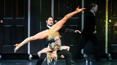 Val Chmerkovskiy hält auf einer Bühne eine Tänzerin im goldenen Kostüm kopfüber mit weit gespreizten Beinen. - Foto: Imago/ZUMA Press Wire