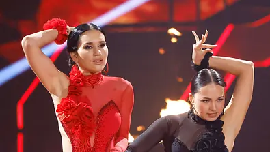 Zwei Tänzerinnen führen eine leidenschaftliche Choreografie auf einer Bühne mit roten Lichteffekten und Flammen im Hintergrund auf. Eine trägt ein rotes, figurbetontes Kleid mit Blumenapplikationen, die andere ein schwarzes Kleid mit transparenten Details. Beide posieren mit erhobenen Armen in einer dramatischen Tanzhaltung. - Foto: IMAGO / Panama Pictures Zwei Tänzerinnen führen eine leidenschaftliche Choreografie auf einer Bühne mit roten Lichteffekten und Flammen im Hintergrund auf. Eine trägt ein rotes, figurbetontes Kleid mit Blumenapplikationen, die andere ein schwarzes Kleid mit transparenten Details. Beide posieren mit erhobenen Armen in einer dramatischen Tanzhaltung. - Foto: IMAGO / Panama Pictures