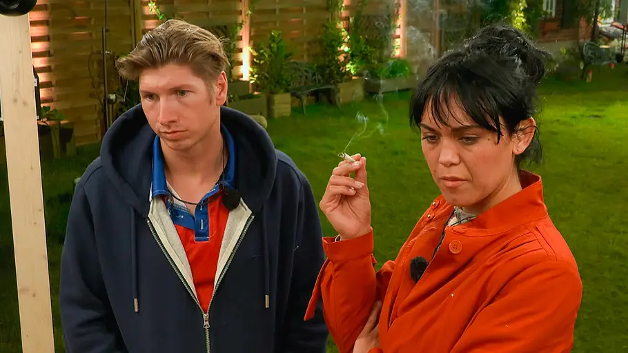 Vanessa Neigert und Giovanni Weisheit Vanessa Neigert hält im orangefarbenen Mantel eine Zigarette. Neben ihr steht Giovanni Weisheit in dunkler Kapuzenjacke im Garten.
