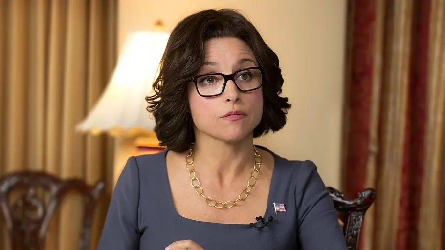 Veep Eine Frau mit dunklem Bob, Brille und grauem Kleid sitzt bei einem Interview oder Gespräch und blickt aufmerksam nach vorne. Sie trägt eine goldene Halskette sowie eine US-Flaggen-Anstecknadel, im Hintergrund sind Vorhänge und eine Stehlampe zu sehen.