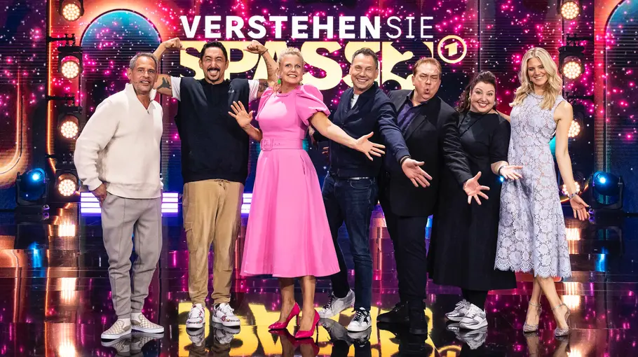 Verstehen sie Spaß? Die Promis und Barbara Schöneberger stehen lachend und mit ausgebreiteten Armen auf einer Bühne vor dem Schriftzug „Verstehen Sie Spaß?“.