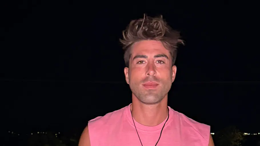 Veysel, 33 Ein Mann in einem rosa ärmellosen Shirt und schwarzer Hose steht nachts vor einem beleuchteten Pool und schaut in die Kamera.